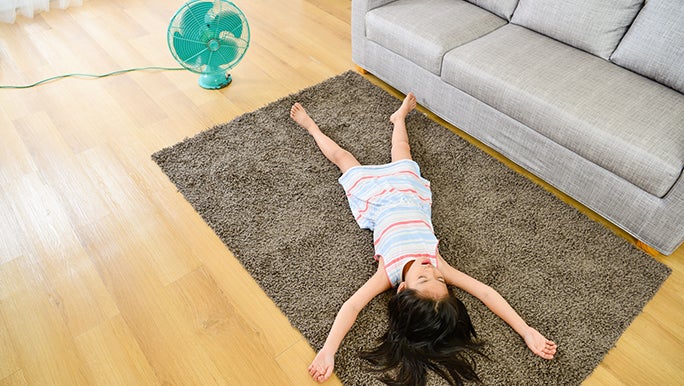 A little girl lays on a rug with her limbs outstretched in the living room. There is a fan on the floor near her.
