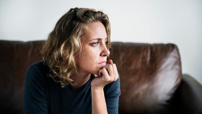 A Caucasian woman is sitting on a brown leather sofa with her chin in her hands, looking stressed and tired, which are symptoms sometimes associated with ‘adrenal fatigue’