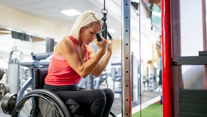Woman in a wheelchair with silver hair using a weights machine. She is doing reps to build muscle. 