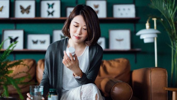  A young Asian woman is sitting on a sofa and reading the label of a bottle of probiotics