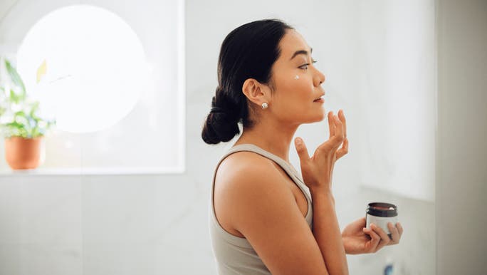 A young Asian woman is holding a jar of face cream and looking at herself in a mirror