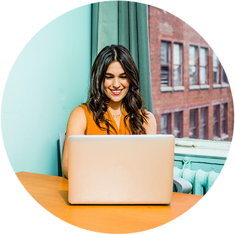 Round photo of a smiling woman with long, dark hair wearing an orange sleeveless top working on her laptop