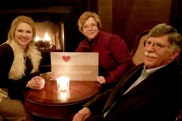 Three ARRT volunteers sitting near a fireplace. One person is holding a placard that says, 