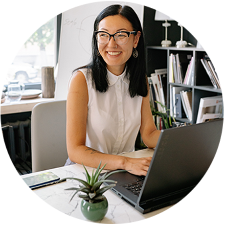 Round photo of a smiling, dark-haired woman with glasses working on a laptop