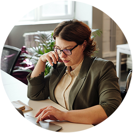 Round photo of a woman with dark hair and glasses, talking on the phone with her right hand, and using a laptop trackpad with her left hand