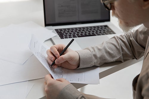 Man completing paper form at a table with a laptop in the background.