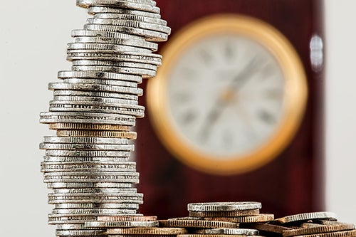 Stack of coins in the foreground with an out-of-focus clock in the background.
