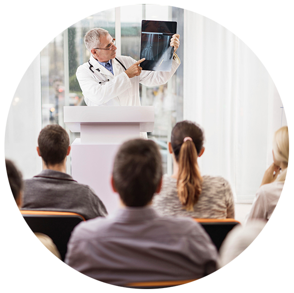 Round photo of a male radiography instructor wearing a lab coat holding up an X-ray in the background; the backs of four students heads in the foreground