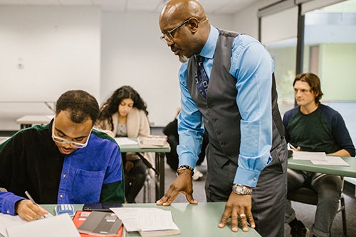A standing instructor helps a seated student with other students seated at desks in the background. 