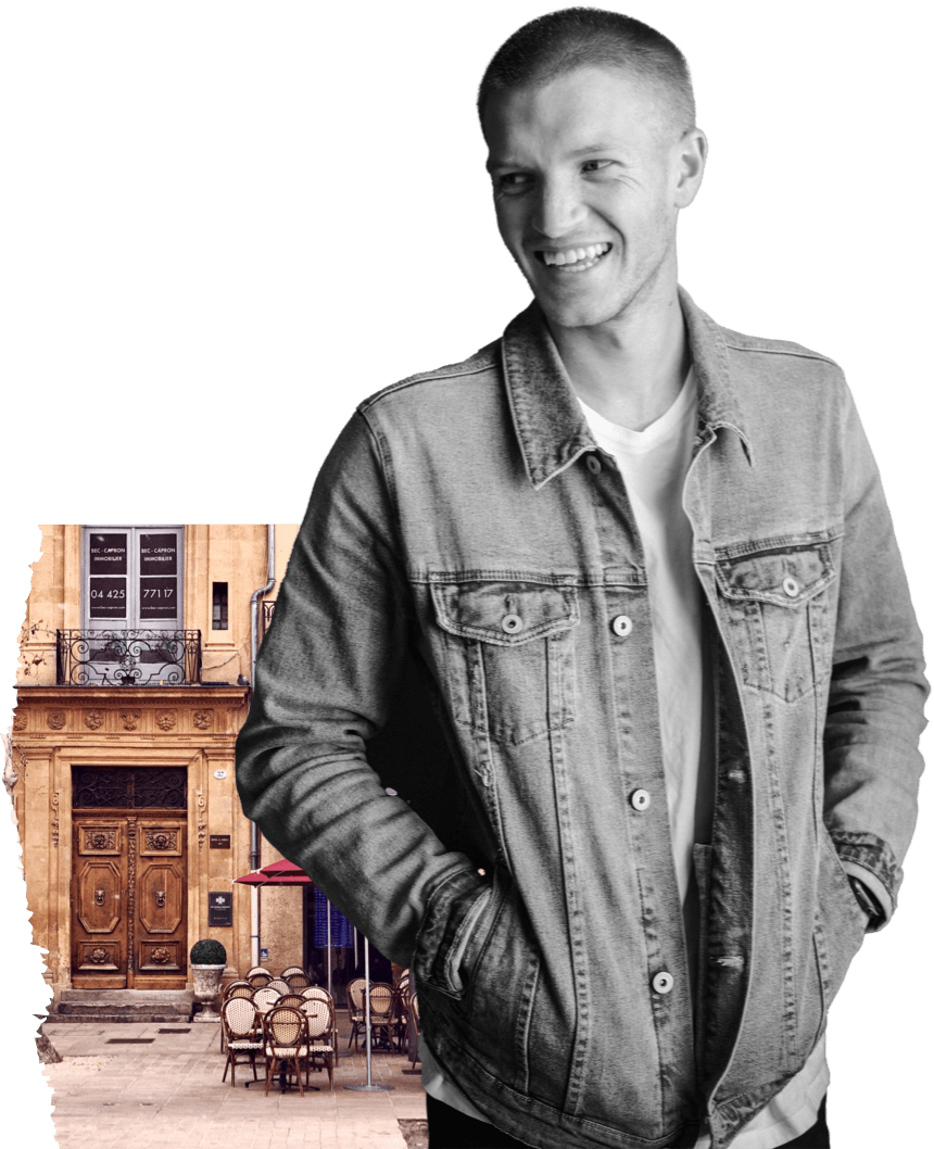 A smiling man in a denim jacket. On his left is the front of an old building with chairs.