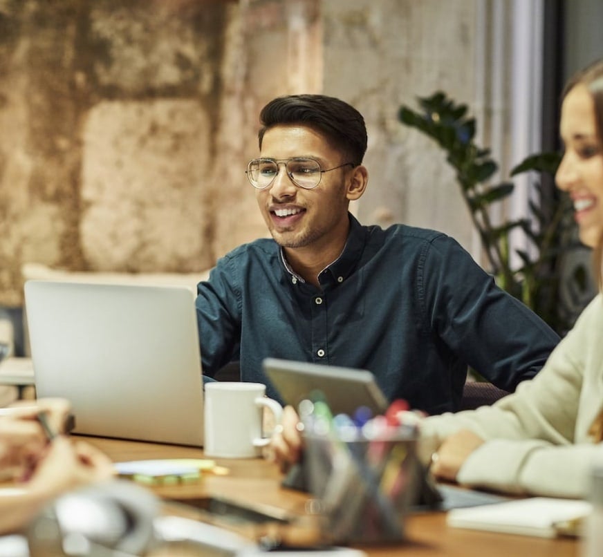 A group of people sitting around a table with laptops & office supplies.