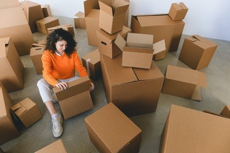 Woman in stylish clothes sitting on floor, packing belongings into cardboard boxes before relocation into new apartment.