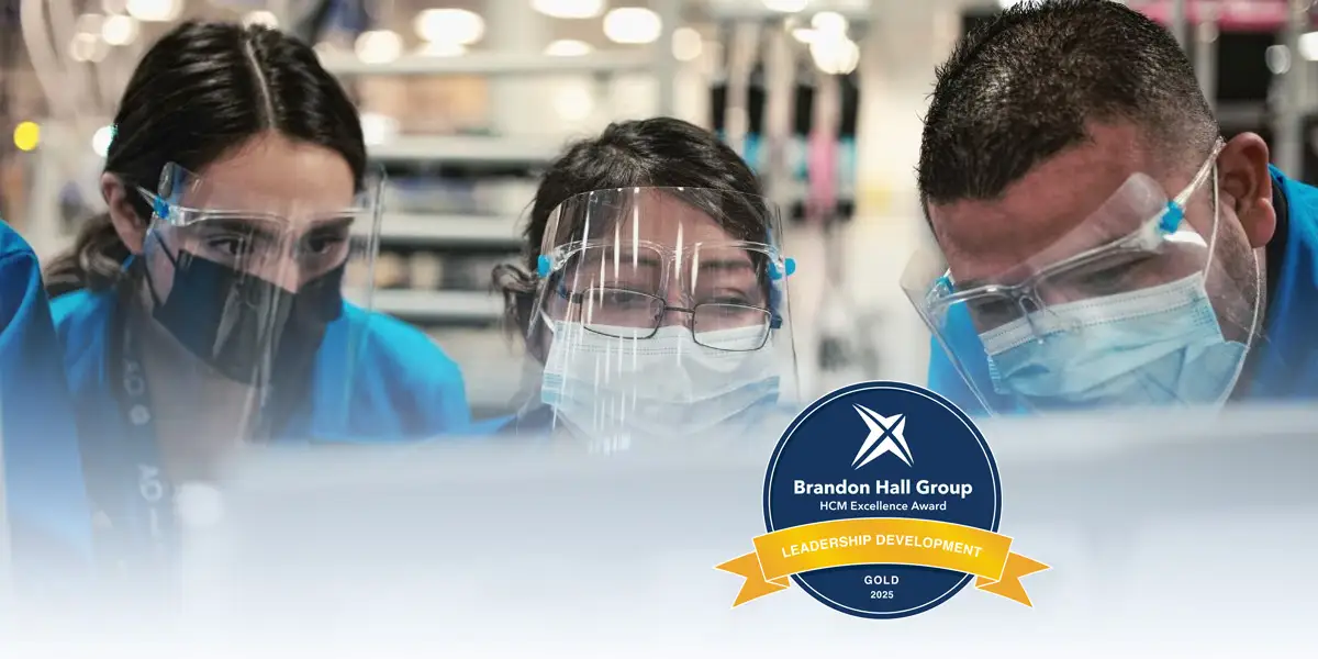 GE HealthCare leaders working together looking at a computer screen, masks and goggles on, and the Brandon Hall HCM Excellence Award 2025 gold badge off in the corner to show the companies recent leadership development program award wins