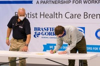 Man bent over signing a paper with another man onlooking