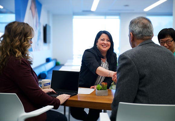three women and a man sat at a table smiling with one woman shaking the mans hand