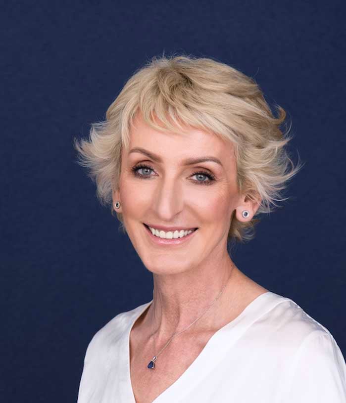 Joanna Bensz, a woman with short blonde hair in a white shirt, wears earrings and a matching necklace on a dark background.
