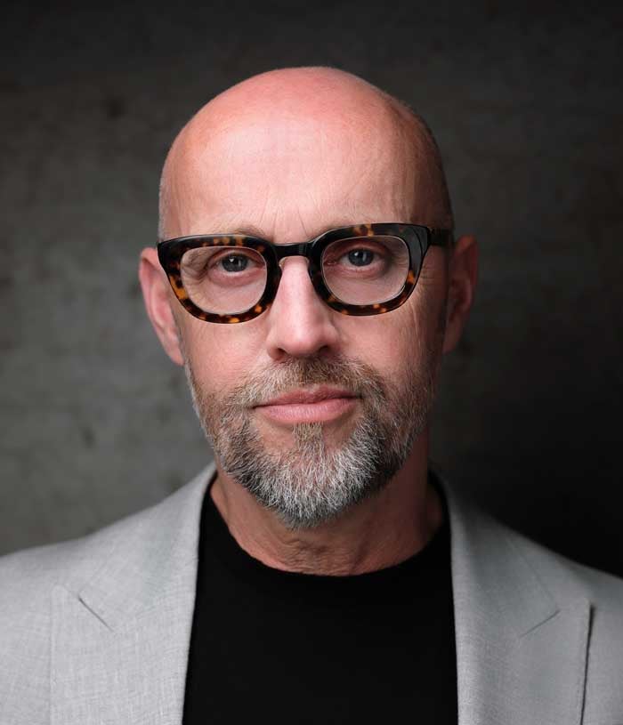 Phil Newman, a bald man with square glasses and a beard, wears a light gray suit and a black shirt.