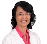 Head-and-shoulders portrait of a person wearing a white medical coat over a bright pink collared shirt, facing forward against a light gray background. The person has short, dark hair styled with volume and is wearing thin, rectangular eyeglasses. The image is evenly lit with a neutral, professional studio appearance.