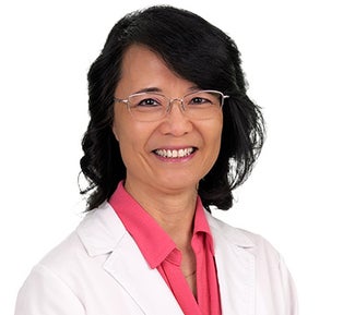 Head-and-shoulders portrait of a person wearing a white medical coat over a bright pink collared shirt, facing forward against a light gray background. The person has short, dark hair styled with volume and is wearing thin, rectangular eyeglasses. The image is evenly lit with a neutral, professional studio appearance.