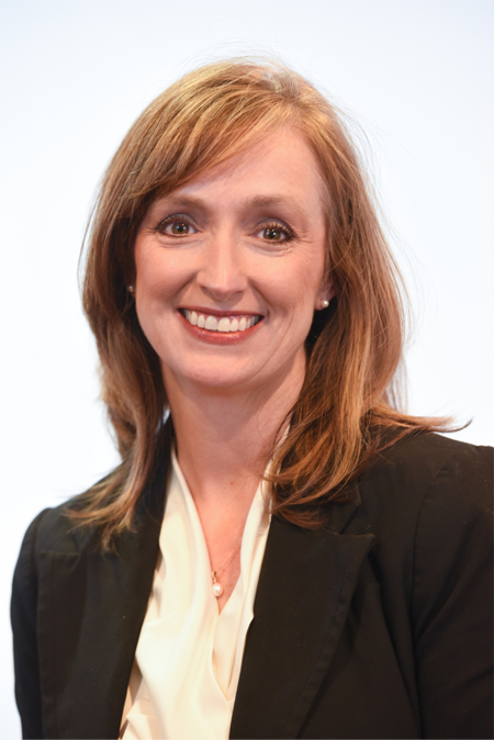 Monica Sherman is pictured, smiling. She has shoulder-length hair that is strawberry blonde and is wearing a black suit jacket and cream top.