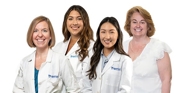 Group portrait of four healthcare professionals standing side by side against a light gray background. Three individuals in the foreground are wearing white medical lab coats with a blue Baptist Health logo on the left chest. One individual is positioned slightly behind the group on the right and is wearing a white blouse. All four individuals are facing the camera. The image is professionally lit with a clean, neutral background and is suitable for marketing, clinical, or healthcare-related materials.