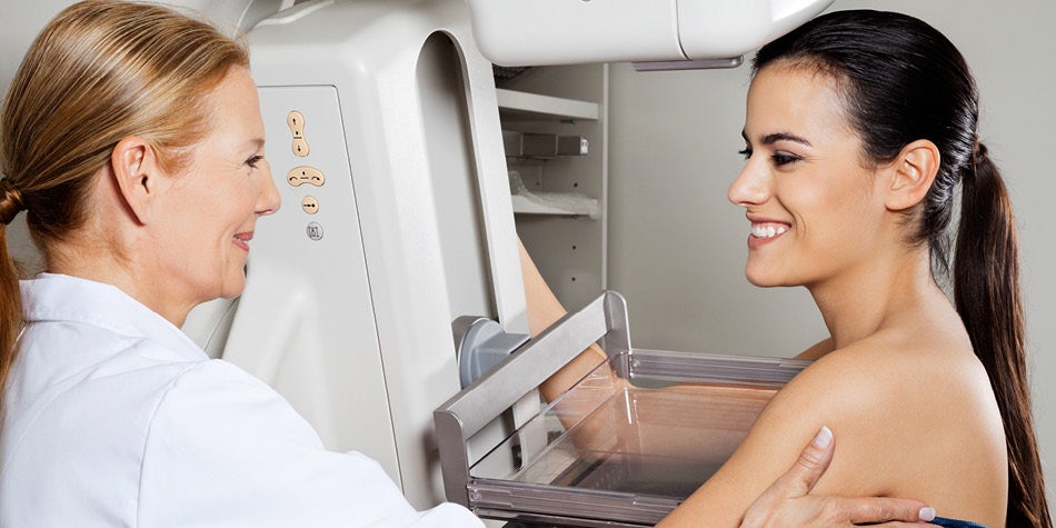 Woman is having a mammogram. Two women are pictured - one using machine and one on the machine for her screening.