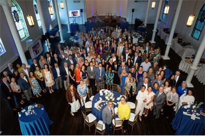 This photos shows a crowd of people with the camera person being up high and looking down. The group is in a dining area with large tables, blue tablecloths, and chairs. The event was in honor of the Baptist Health Care Foundation's 50th anniversary.