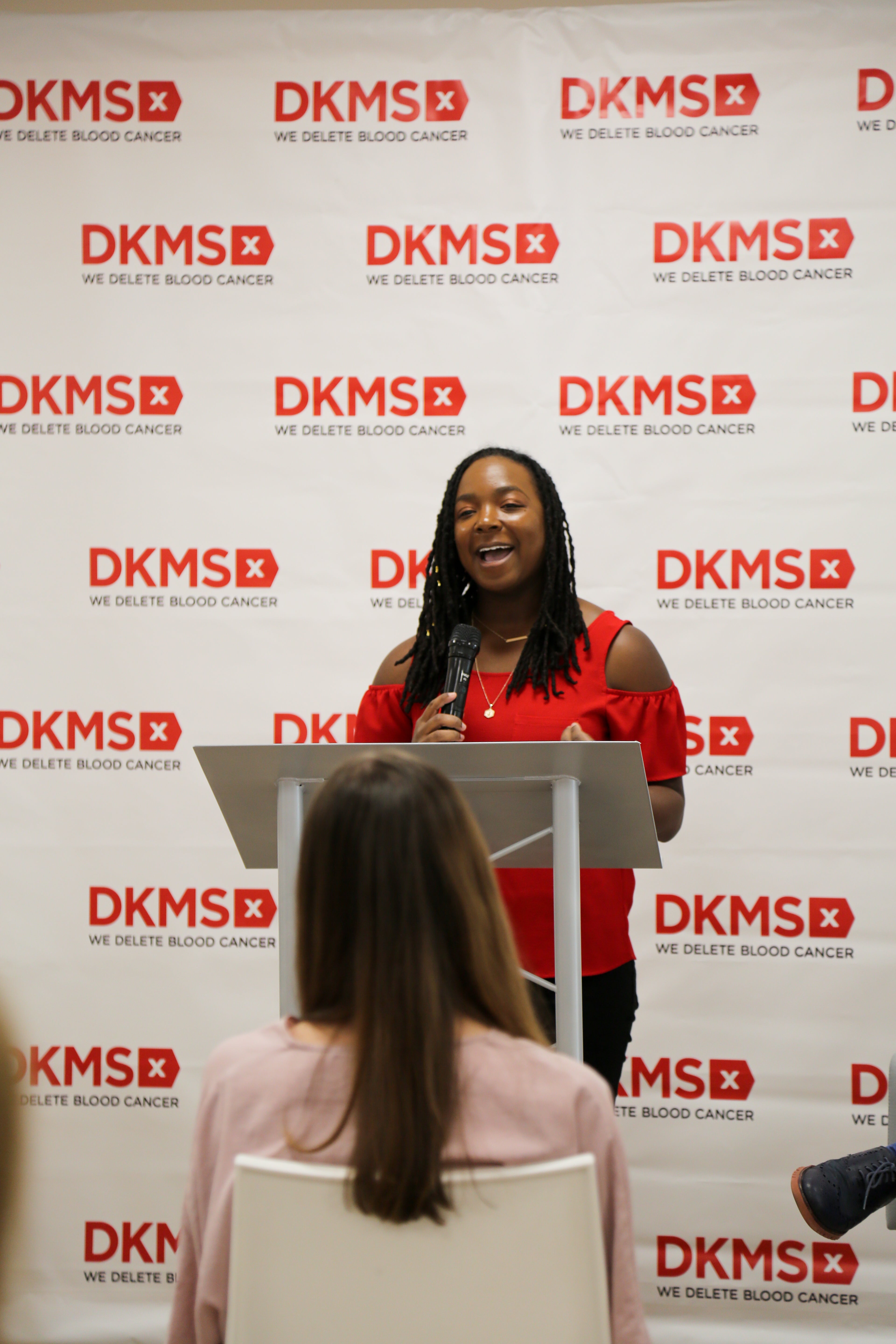 DKMS Stem Cell Donor, Jasmine Deberry Thompson, standing at the podium giving a speech in front of a DKMS banner. 