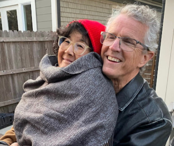 Mama G is curled up in a gray blanket while wearing a red hat. She is sitting on her husband's lap. Both are smiling. 