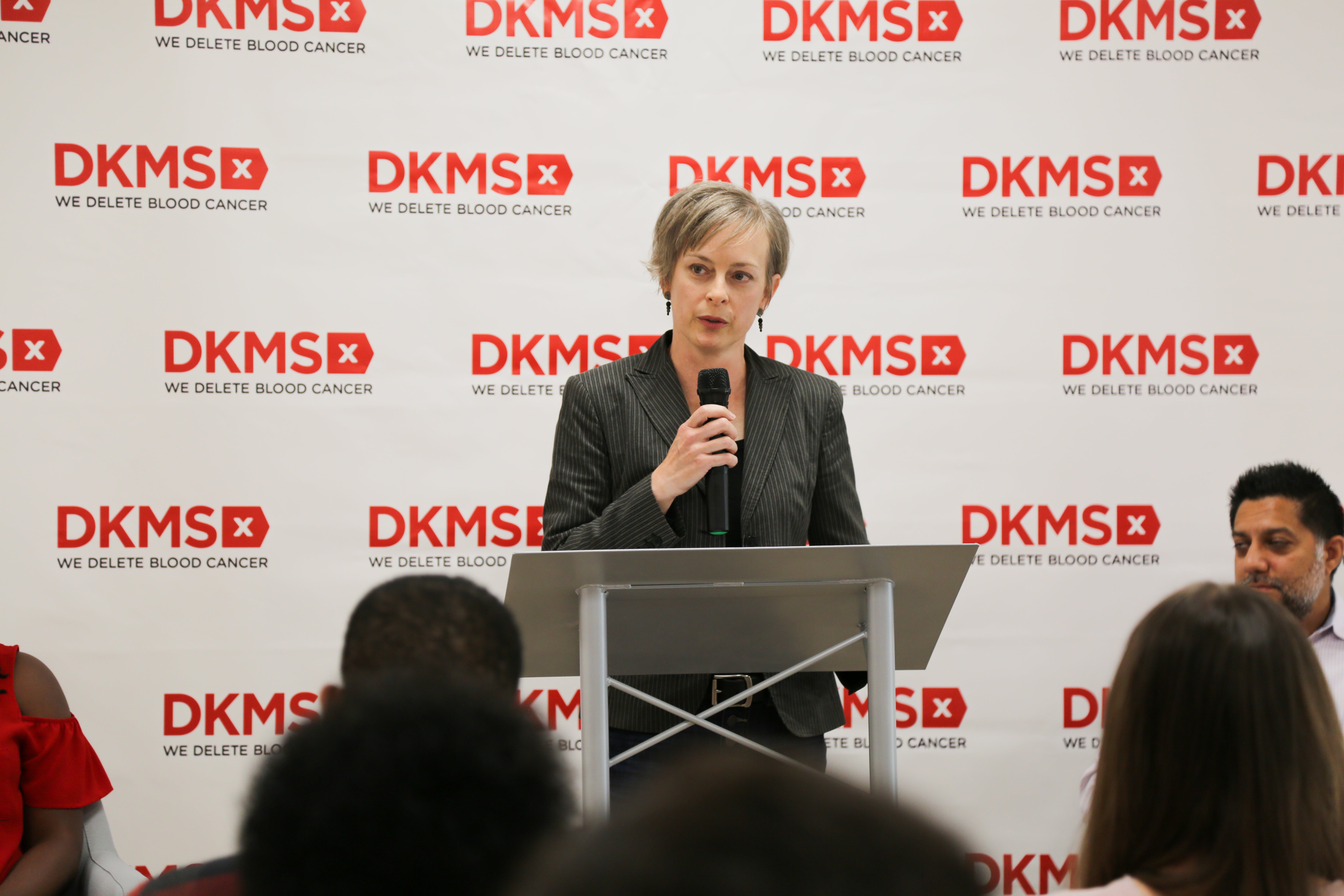 Stem cell transplant recipient, Kristi Martin, standing at the podium speaking in front of a DKMS banner 