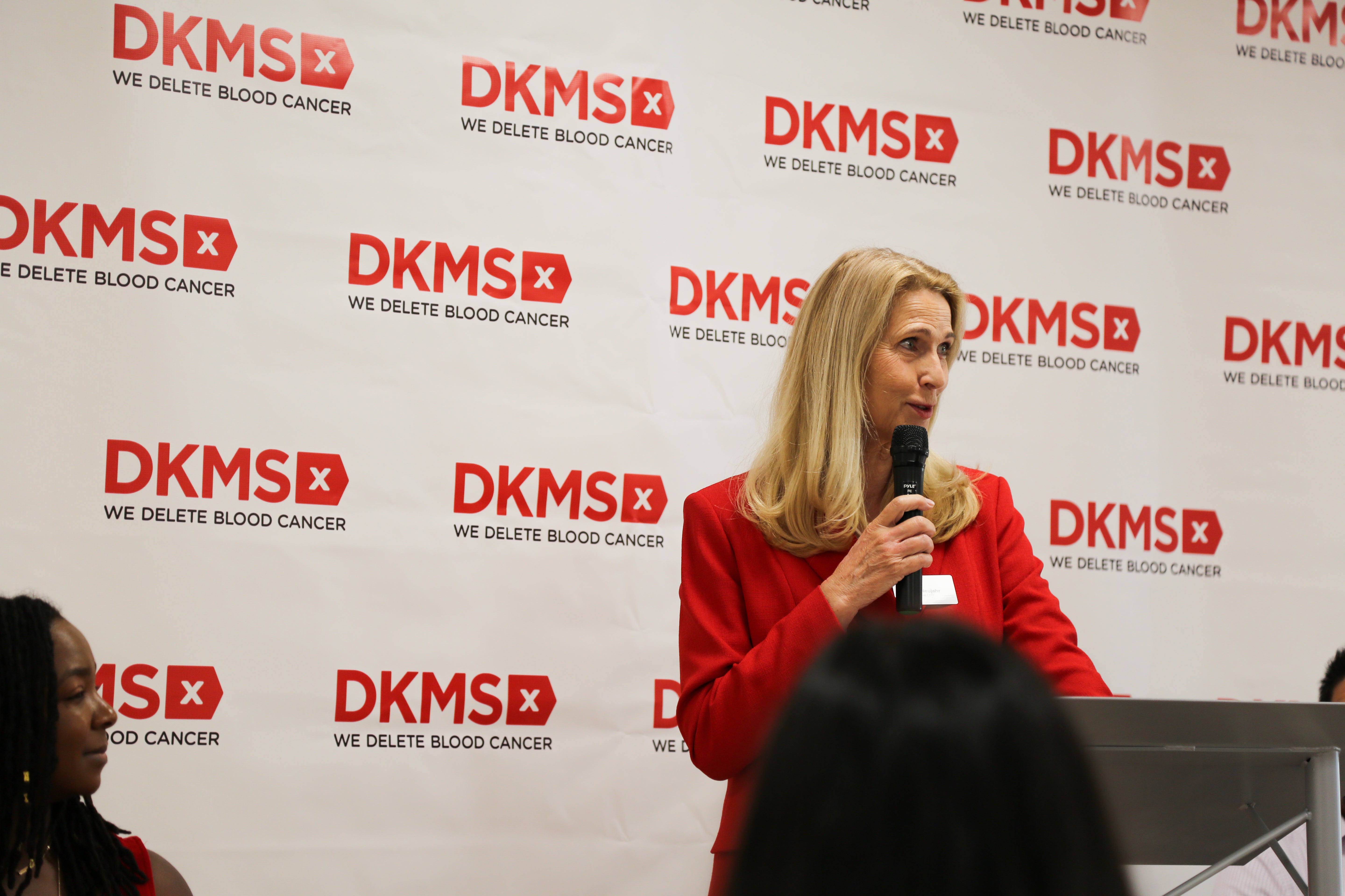 Dr. Elke Neujahr, DKMS Group Global CEO, stands in front of the podium speaking with the DKMS banner in the background.