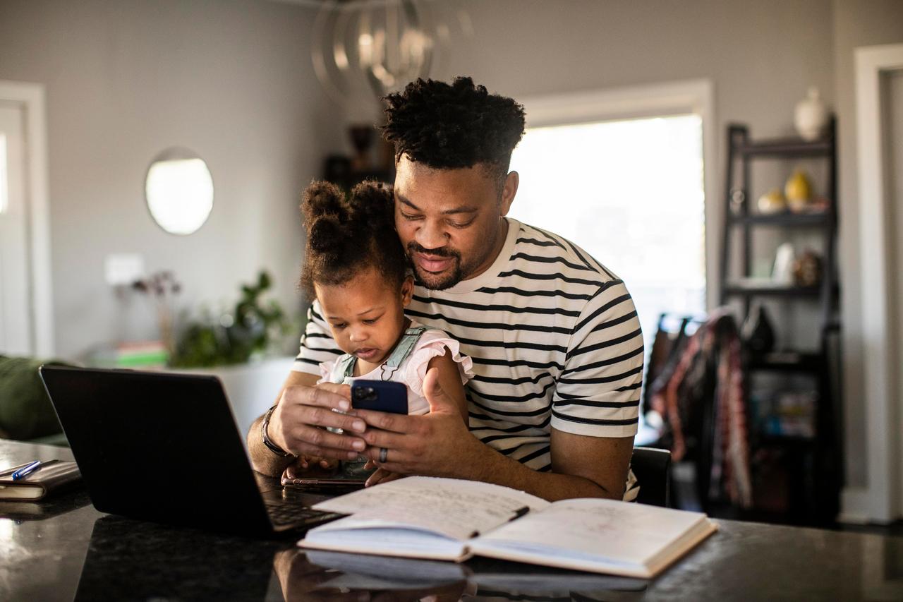 Father working on laptop and phone while holding toddler
