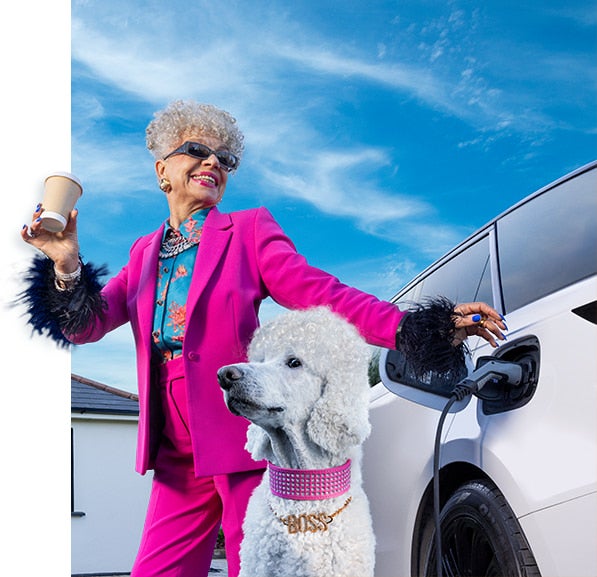 Person in a bright magenta suit plugging in a white electric car charger while holding a coffee cup, with a white poodle wearing a pink rhinestone “BOSS” collar nearby under a blue sky.