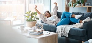 Person stretching on a grey sofa in a bright modern living room with indoor plants and books on a coffee table.