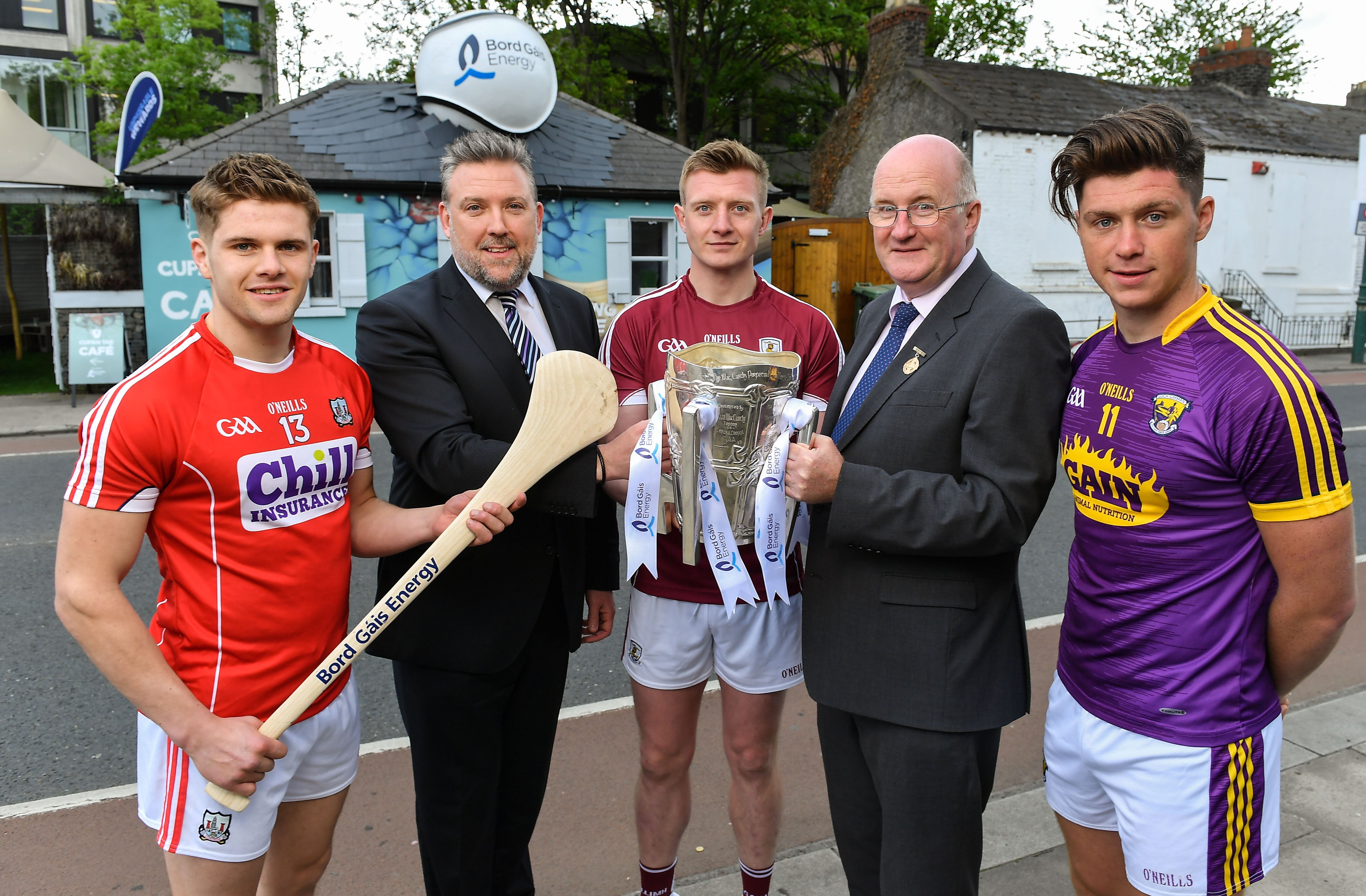  5 men standing outside a cafe: 3 county hurlers, one holding a hurl, one holding a trophy; 2 men in suits.