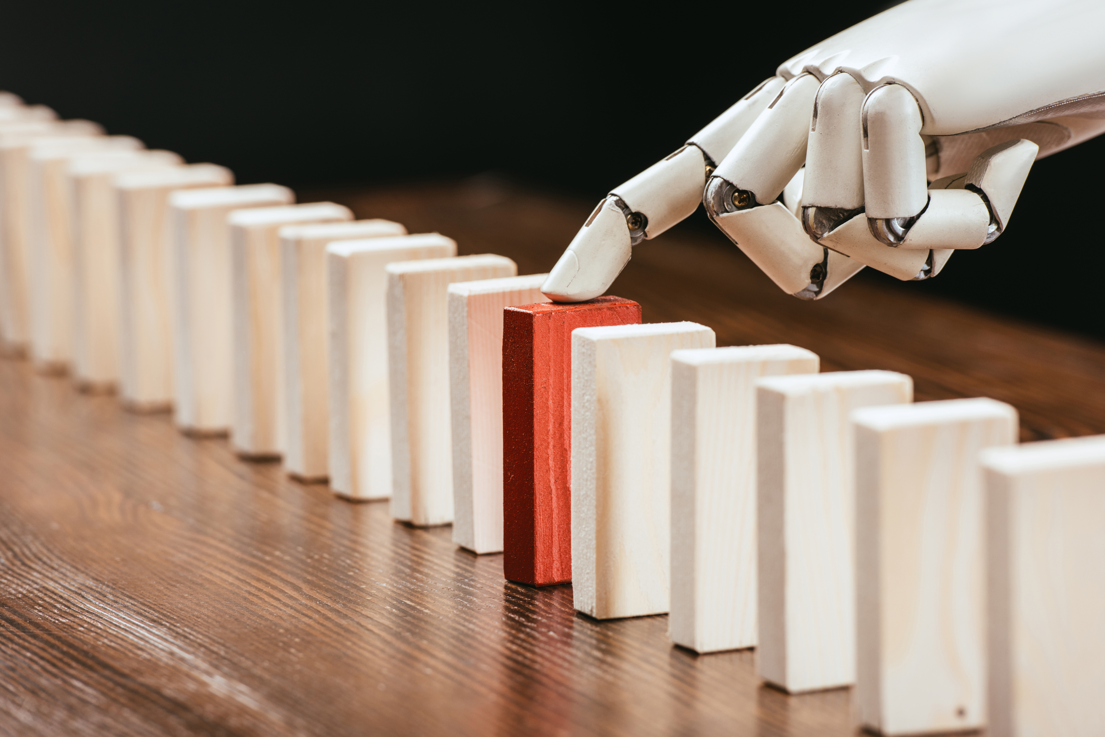 A white robotic hand pushes a red domino in a line of light wooden dominoes. They all stand on a grained wooden table. The background is dark. The close-up shot highlights the contrast in color between the red domino and the other dominoes.