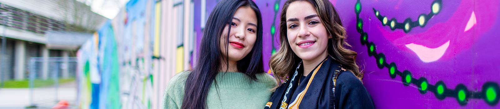 A pair of students smiling in front of colourful graffiti.