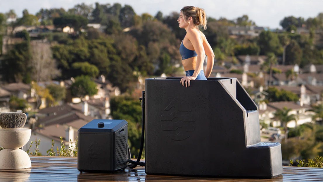 A woman in athletic attire standing in an Ice Barrel cold plunge tub with a play button overlaid on the image