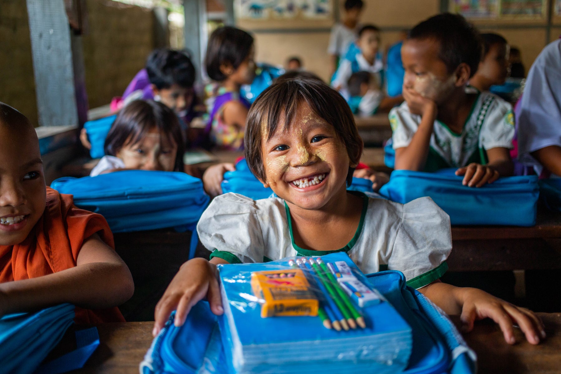 A smiling young girl from Myanmar sits in a classroom with her UNICEF backpack and stationary kit. 