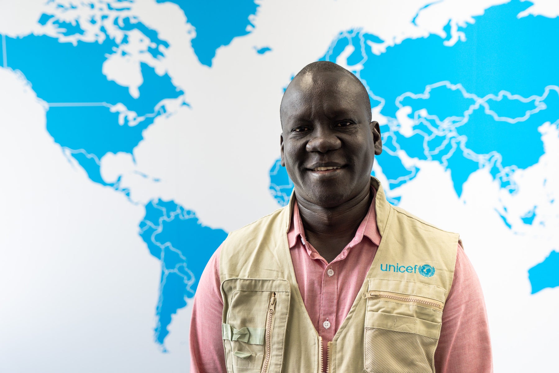 Bismarck Swangin, wearing a beige UNICEF vest and pink button up shirt, poses for a photo behind a wall featuring a blue map of the world.