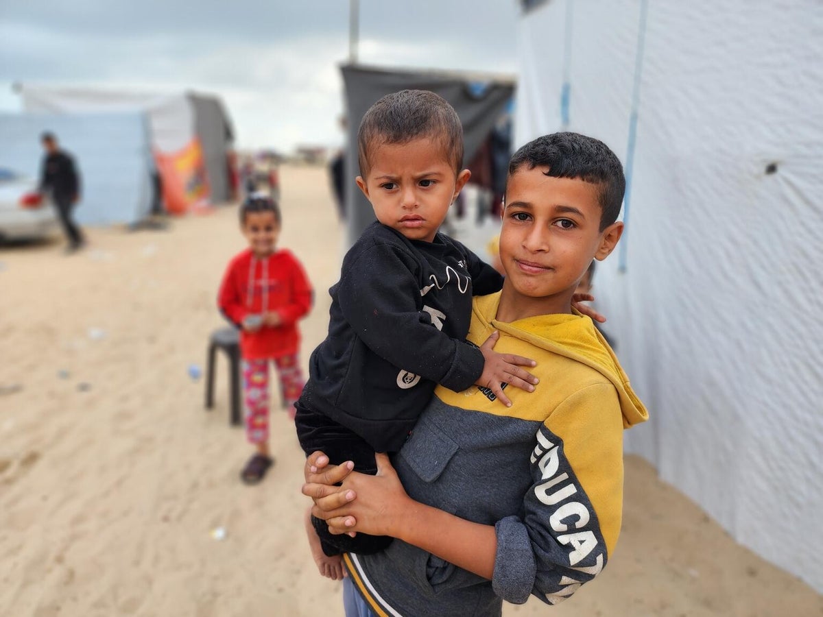 At 14, Mohammed holds his two-year-old brother in front of their temporary shelter in the Gaza Strip. By May 2024, Mohammed and his family had been displaced four times.