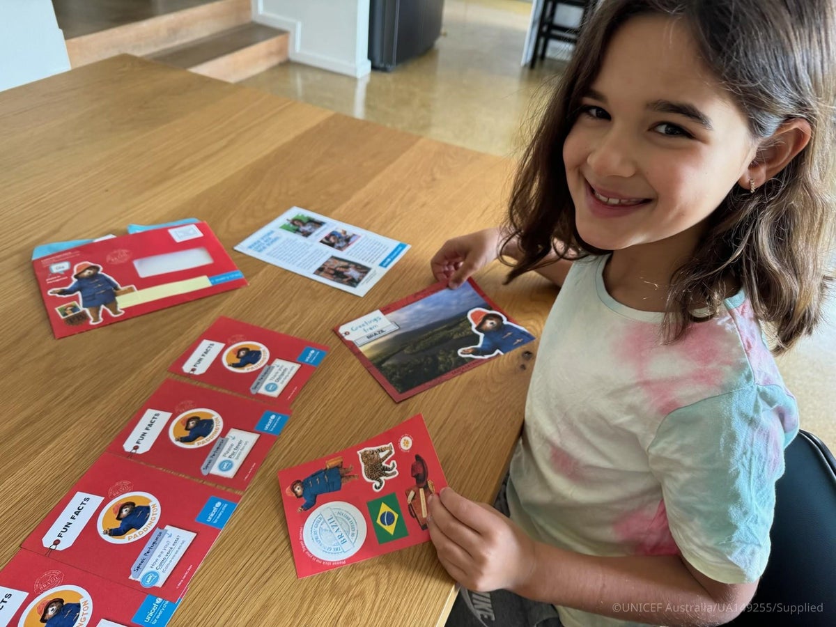 A girl sitting at a table, holding a Paddington’s Postcards card, with postcards, stickers and activity cards spread out in front of her.