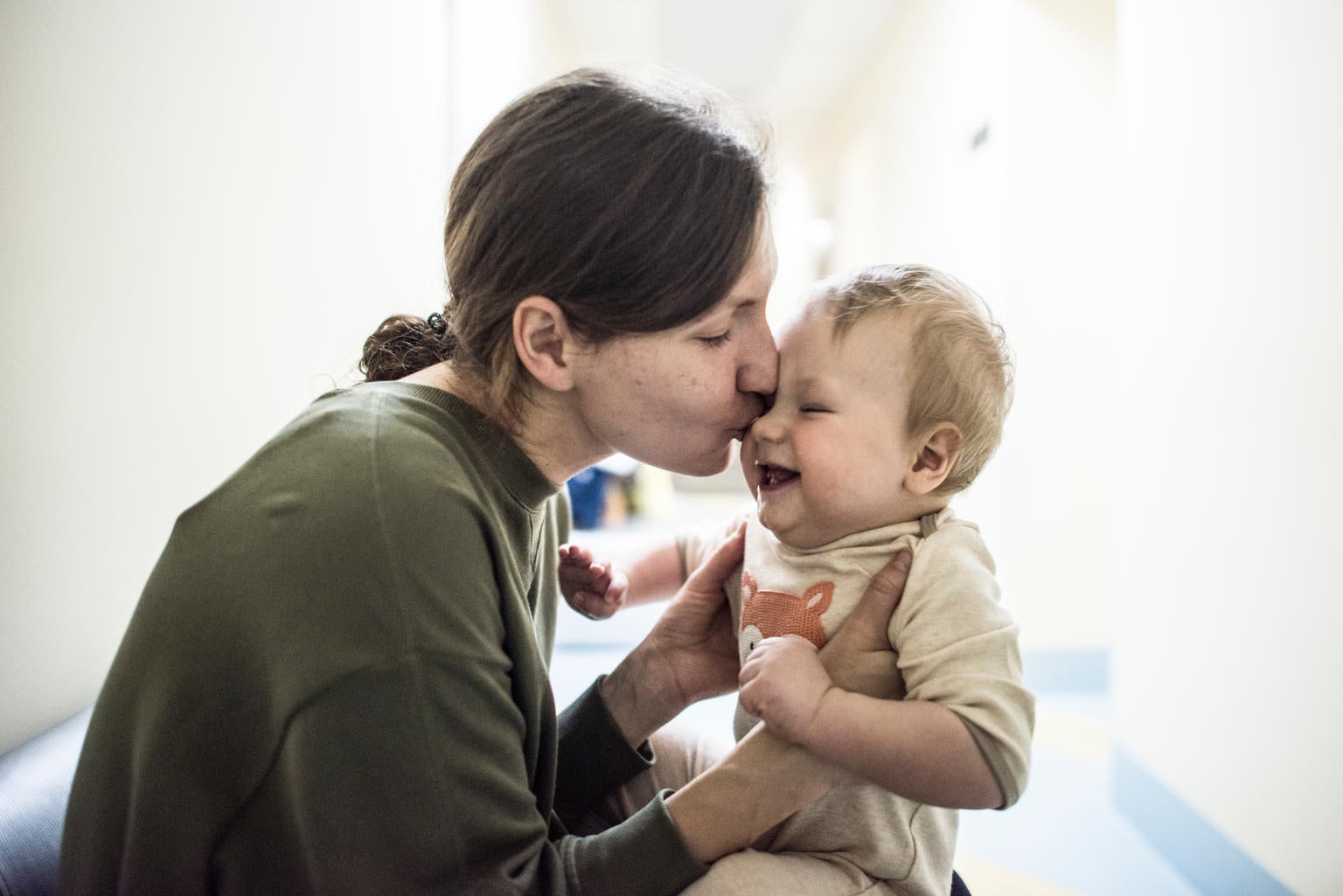 A woman is holding a baby and is kissing them on the cheek. The baby is smiling.