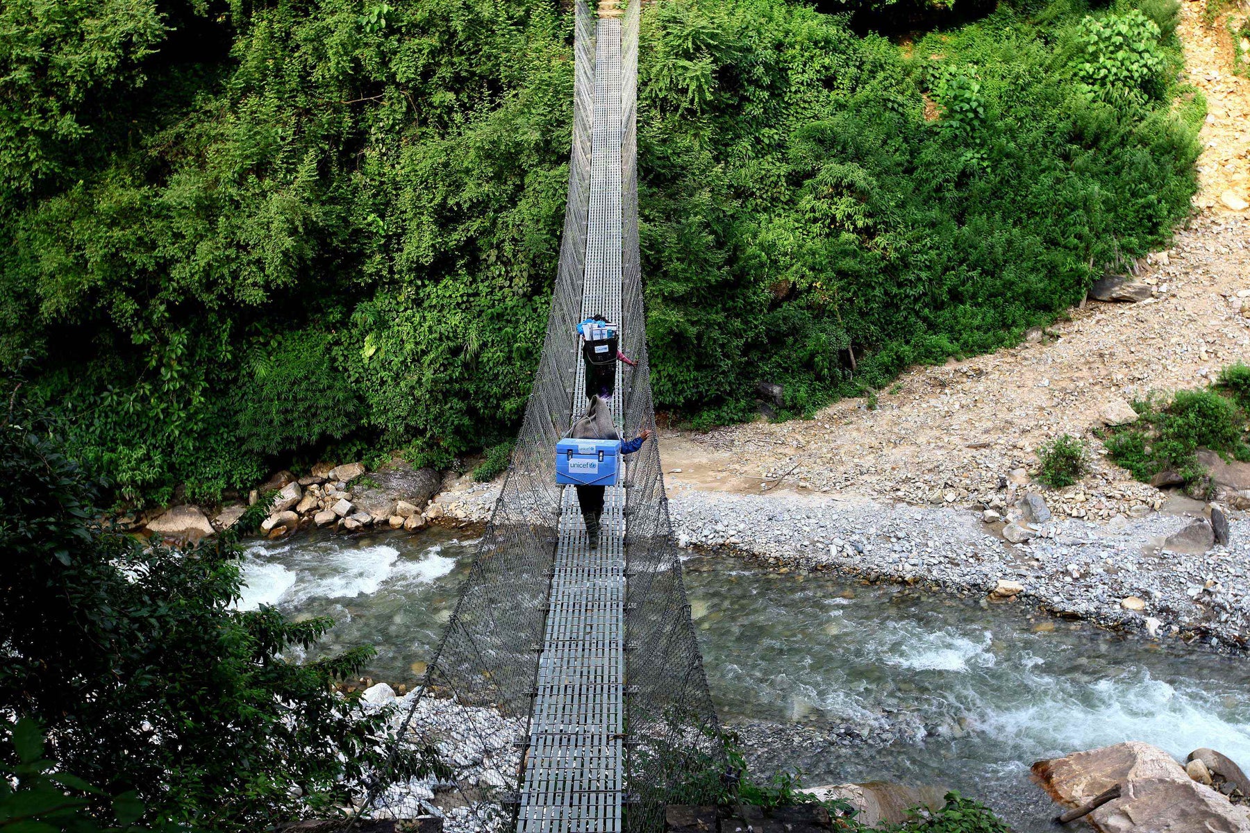 Two people crossing a suspension bridge over a river. They have cooler boxes on their backs