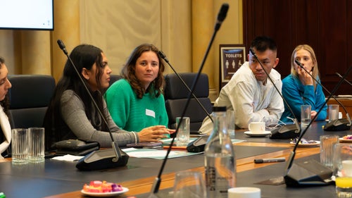 A group of young people sitting around a table watch on as a young woman speaks.