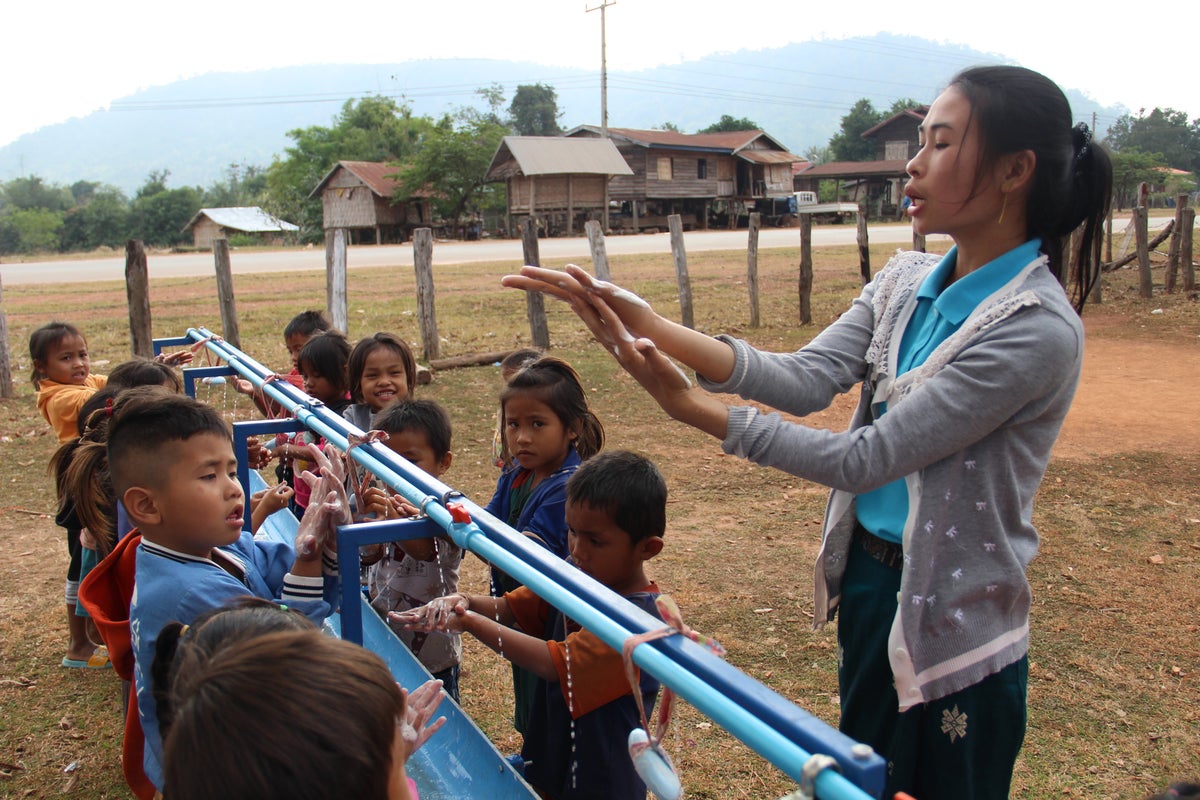 A woman is demonstrating how to wash hands to a group of young children. The children are using outdoor washing facilities.