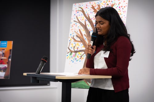 A young woman holds a microphone as she speaks at a lectern. 