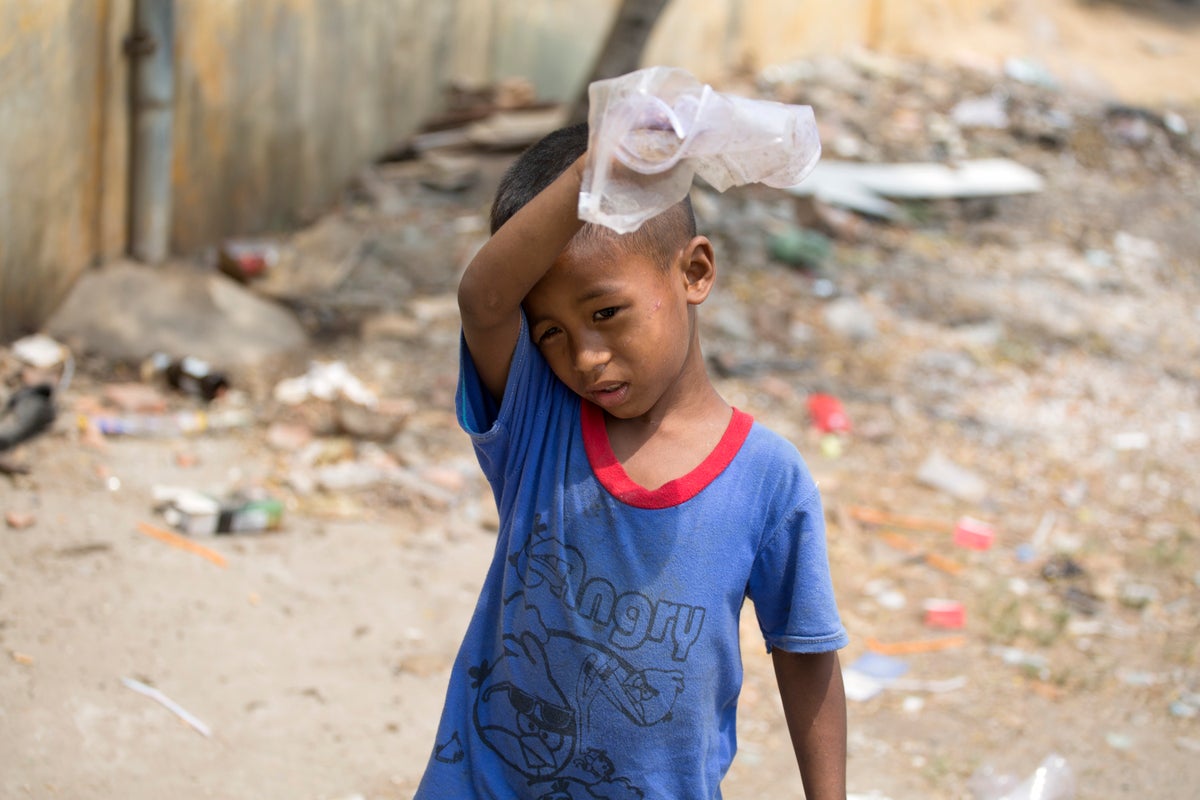6 year-old in blue shirt boy helps his mum collect rubbish to sell in Phnom Penh. He carries a plastic bag and is wiping his forehead.