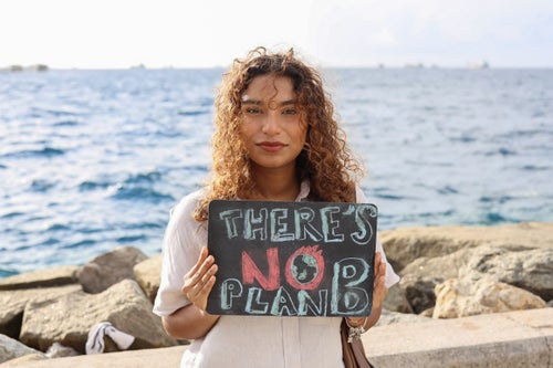 A young woman stands in front of the ocean in the Maldives holding up a sign saying, ‘There’s no Plan B’. 