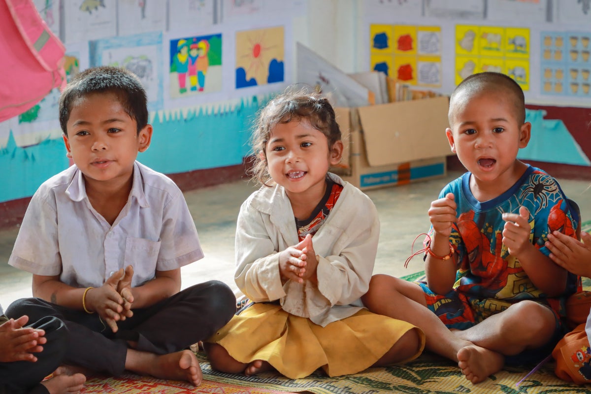 Three children sitting with their legs crossed on the ground clap their hands in their classroom in Lao PDR.
